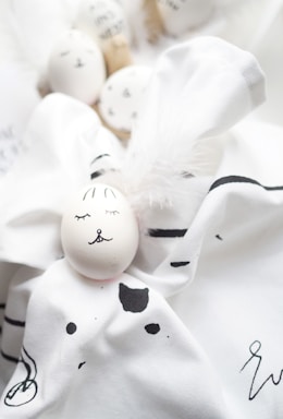 Several white eggs are artistically decorated with simple facial features and placed on a soft, white fabric with black patterns. The arrangement includes fluffy white feathers, contributing to a gentle and whimsical aesthetic.