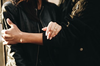 A close-up of two people wearing dark jackets, one person has their arm around the other. The focus is on the hand of one person resting on the other's arm, with a visible ring on the finger.