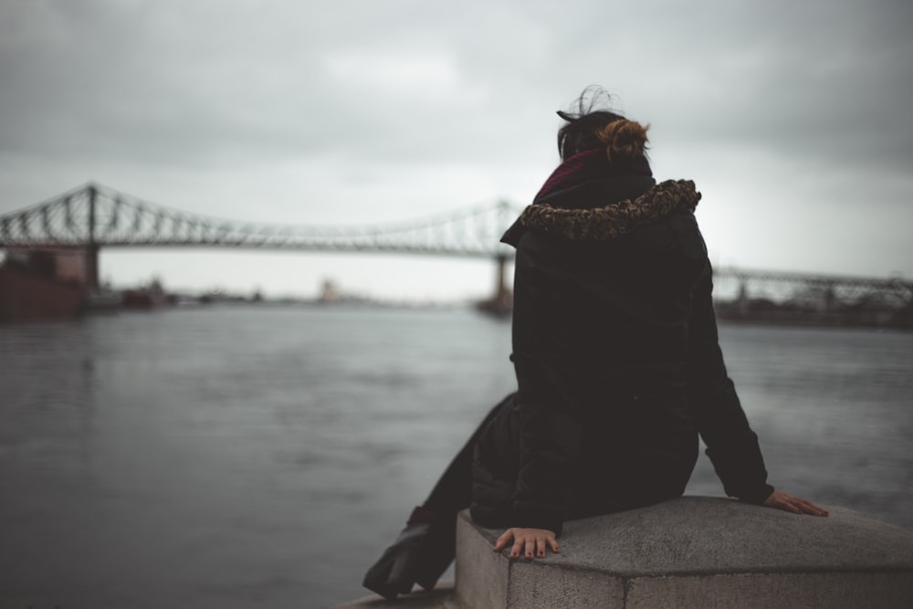 woman sitting on gray concrete table near body of water overlooking bridge