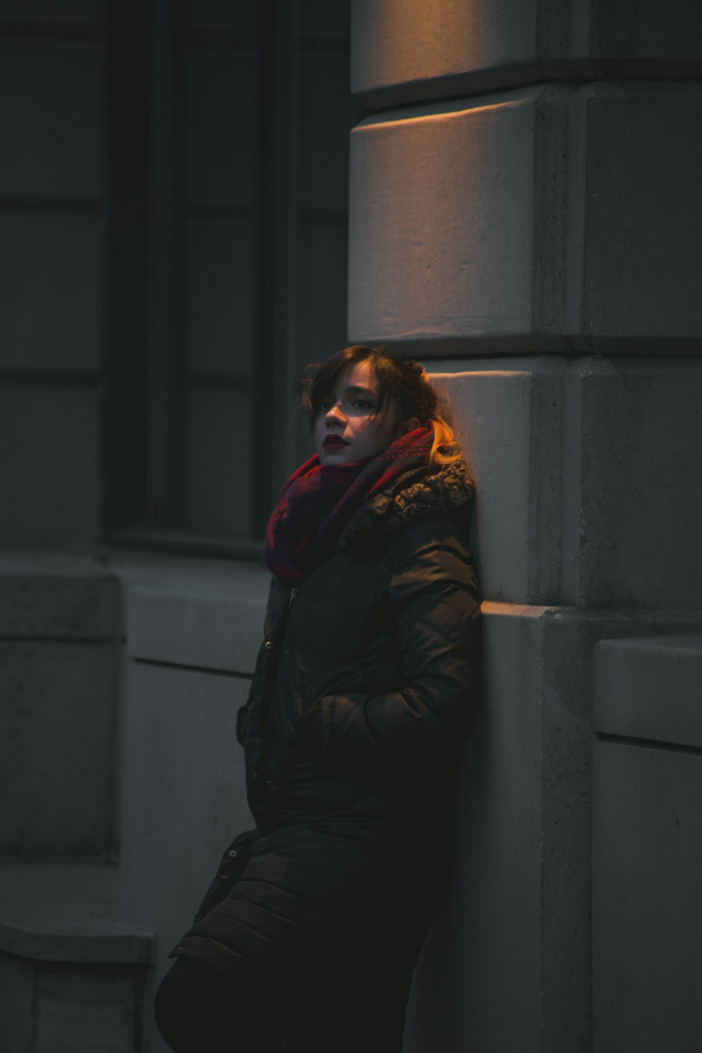 woman in black bubble jacket leaning on gray wall during nighttime