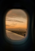 Looking through an airplane window, a view of another plane on the tarmac is visible during sunset. The sky is filled with soft, warm hues of orange and yellow, casting a serene glow over the scene. A Delta airplane is positioned prominently in the distance.