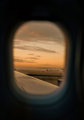 Looking through an airplane window, a view of another plane on the tarmac is visible during sunset. The sky is filled with soft, warm hues of orange and yellow, casting a serene glow over the scene. A Delta airplane is positioned prominently in the distance.