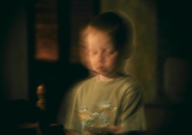 A blurred image of a young child with short hair, wearing a t-shirt featuring cartoon characters. The background is dark, giving a dreamlike or otherworldly effect.