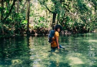 person in orange top wearing backpack walking on body of water in forest during daytime