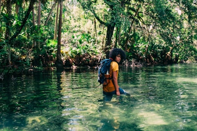 person in orange top wearing backpack walking on body of water in forest during daytime