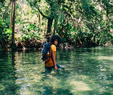 person in orange top wearing backpack walking on body of water in forest during daytime