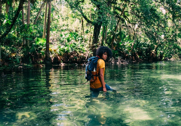 person in orange top wearing backpack walking on body of water in forest during daytime