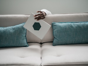 A light-colored sofa with two teal cushions is seen, along with a hand holding a white rectangular object featuring a hexagonal design situated between the cushions.
