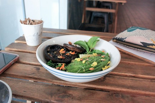 A wooden table with a bowl of fresh salad featuring sliced avocado, grilled mushrooms, mint leaves, and sprinkled seeds and flower petals. Nearby are a paper cup with wooden stirrers, a smartphone, and a colorful patterned pouch.
