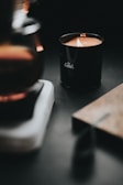 A lit candle in a dark, cozy setting, with the words 'the Goods' written on the black container. The surrounding area includes a blurred surface and warm lighting, creating a tranquil and intimate atmosphere.