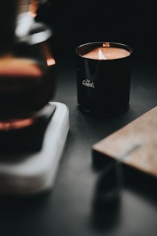 A lit candle in a dark, cozy setting, with the words 'the Goods' written on the black container. The surrounding area includes a blurred surface and warm lighting, creating a tranquil and intimate atmosphere.