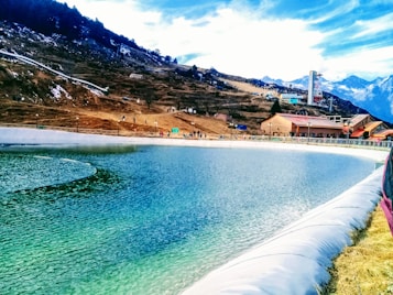 A clear turquoise water body is surrounded by a snow-kissed mountain landscape. Buildings and small structures are visible near the water, with some people walking along the paths. The sky is partly cloudy and the scene is set against a backdrop of majestic peaks.
