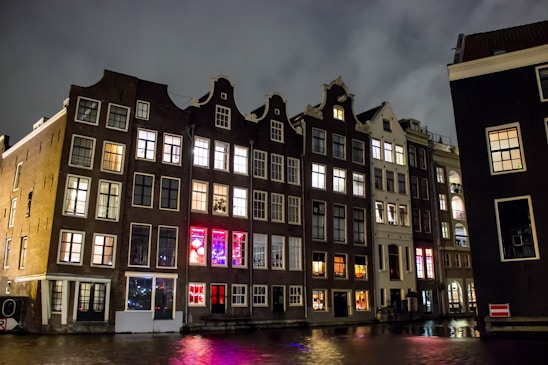 Tall, narrow buildings with large windows are illuminated at night, reflected on a canal. The architecture features gabled roofs, typical of Dutch style. Some windows display colorful lights, adding a vibrant touch against the cloudy night sky.