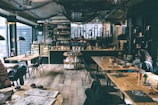 A cozy coffee shop interior with wooden floors and tables, featuring a well-organized bar with various bottles and a chalkboard menu. Soft lighting and a few patrons sitting and enjoying their beverages contribute to a relaxed atmosphere. The decor includes shelves with items and a circular display full of products.