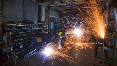 A dimly lit workshop with two workers using metal grinders, creating bright, cascading sparks that illuminate the area. Shelves filled with tools and equipment can be seen on the left side, with a cluttered, industrial environment surrounding the workers.
