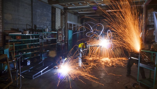 A dimly lit workshop with two workers using metal grinders, creating bright, cascading sparks that illuminate the area. Shelves filled with tools and equipment can be seen on the left side, with a cluttered, industrial environment surrounding the workers.