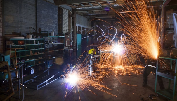 A dimly lit workshop with two workers using metal grinders, creating bright, cascading sparks that illuminate the area. Shelves filled with tools and equipment can be seen on the left side, with a cluttered, industrial environment surrounding the workers.