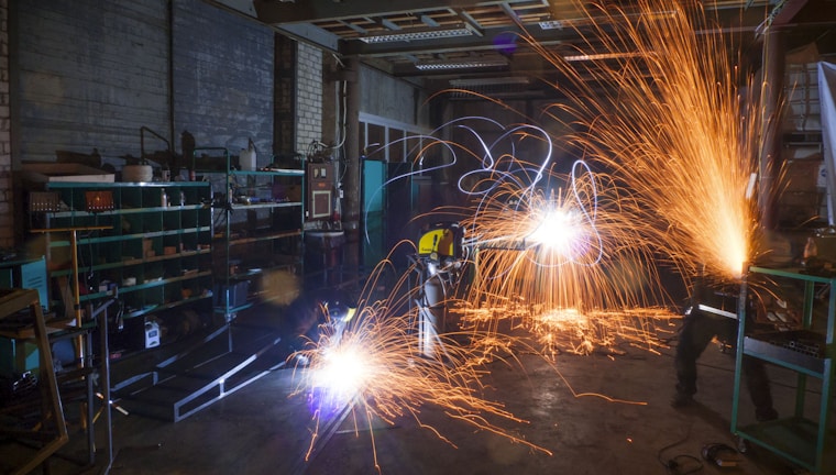 A dimly lit workshop with two workers using metal grinders, creating bright, cascading sparks that illuminate the area. Shelves filled with tools and equipment can be seen on the left side, with a cluttered, industrial environment surrounding the workers.