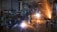 A dimly lit workshop with two workers using metal grinders, creating bright, cascading sparks that illuminate the area. Shelves filled with tools and equipment can be seen on the left side, with a cluttered, industrial environment surrounding the workers.