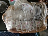 A set of white porcelain teacups and saucers with floral patterns, wrapped in clear plastic for protection. The items are arranged in a wicker basket, showcasing elegant floral designs including pink and yellow flowers.