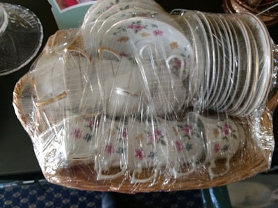 A set of white porcelain teacups and saucers with floral patterns, wrapped in clear plastic for protection. The items are arranged in a wicker basket, showcasing elegant floral designs including pink and yellow flowers.