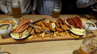 A large wooden board is filled with various meats and side dishes. There are different types of sausages, fried chicken cutlets, and potato wedges. Small bowls hold mustard and a creamy soup, and a plate with shredded carrots and greens is also present. Multiple mugs of beer are on the table, and three people are partially visible.