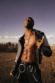 A person stands in an open outdoor area with a clear sky. They wear a studded leather jacket, chains, and accessories, creating a fashionable and edgy appearance.