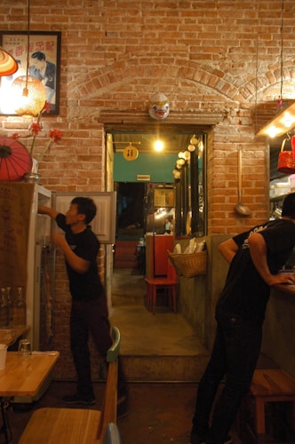 An interior scene of a cozy, rustic restaurant with exposed brick walls and warm lighting. Two individuals in black shirts are seen, one opening a refrigerator and the other working near a counter. A wooden table with chairs is in the foreground. Decorative elements include a colorful hanging lamp, a traditional mask mounted above a doorway, and a framed poster. The doorway leads to another dimly lit area with red chairs.
