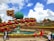 The scene features a vibrant botanical display with numerous multi-layered flower arrangements in red, yellow, and green, encircling a large green spherical structure. People are walking and taking pictures, with a clear blue sky and white clouds in the background.