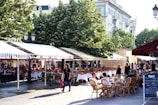 An outdoor cafe features numerous tables with white tablecloths and red napkins set under striped canopy awnings. People are seated at wicker chairs, enjoying the ambiance. In the background, lush green trees and historical buildings are visible. The setting is lively with a pedestrian carrying a red shopping bag strolling by.