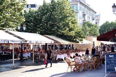 An outdoor cafe features numerous tables with white tablecloths and red napkins set under striped canopy awnings. People are seated at wicker chairs, enjoying the ambiance. In the background, lush green trees and historical buildings are visible. The setting is lively with a pedestrian carrying a red shopping bag strolling by.