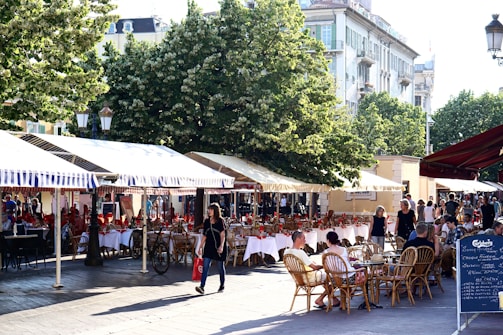An outdoor cafe features numerous tables with white tablecloths and red napkins set under striped canopy awnings. People are seated at wicker chairs, enjoying the ambiance. In the background, lush green trees and historical buildings are visible. The setting is lively with a pedestrian carrying a red shopping bag strolling by.