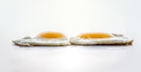 Two sunny-side-up eggs are placed side by side on a flat surface. The whites are cooked through, with a slight crispiness around the edges, while the yolks remain bright and runny. The background is softly blurred, emphasizing the texture and color of the eggs.