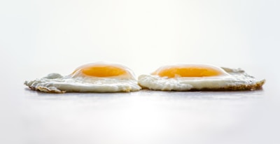 Two sunny-side-up eggs are placed side by side on a flat surface. The whites are cooked through, with a slight crispiness around the edges, while the yolks remain bright and runny. The background is softly blurred, emphasizing the texture and color of the eggs.