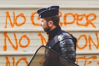 A person in riot gear stands in front of a wall with orange graffiti that reads 'NO ORDER NO'. The individual is wearing a dark uniform and riot helmet, carrying a transparent shield, suggesting preparation for crowd control or protest situations.