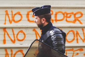 A person in riot gear stands in front of a wall with orange graffiti that reads 'NO ORDER NO'. The individual is wearing a dark uniform and riot helmet, carrying a transparent shield, suggesting preparation for crowd control or protest situations.