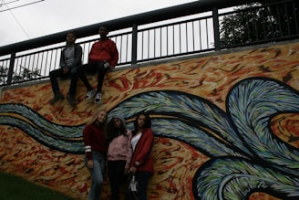 A group of four young people pose against a vibrant mural with abstract, swirling patterns. Two individuals sit on the ledge above the mural, while two others stand in front of it. The mural features wavy designs in shades of blue, green, and orange with a textured background.