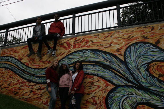 A group of four young people pose against a vibrant mural with abstract, swirling patterns. Two individuals sit on the ledge above the mural, while two others stand in front of it. The mural features wavy designs in shades of blue, green, and orange with a textured background.