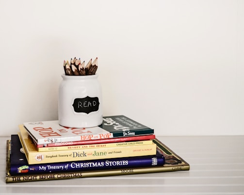 A stack of children's books is neatly arranged on a white surface. On top of the stack is a white jar labeled 'READ' filled with twig-like colored pencils. The books include titles like Hop on Pop by Dr. Seuss, Pinocchio, and Beauty and the Beast.