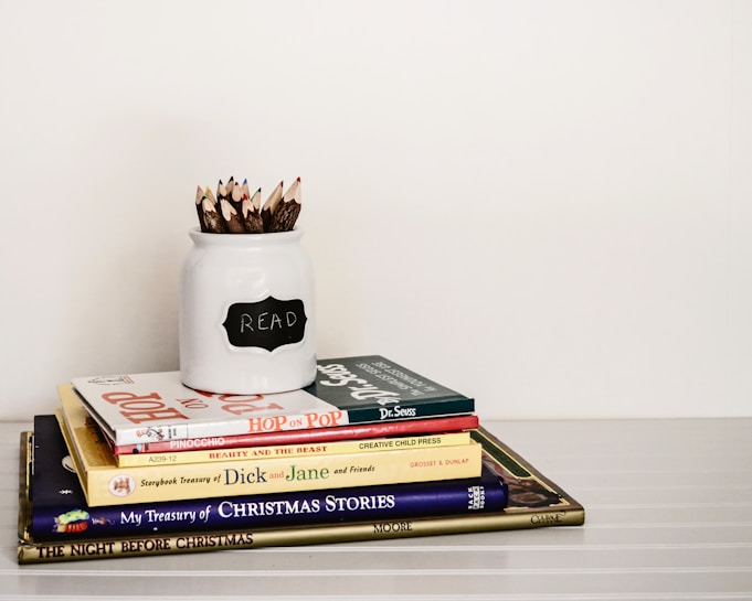A stack of children's books is neatly arranged on a white surface. On top of the stack is a white jar labeled 'READ' filled with twig-like colored pencils. The books include titles like Hop on Pop by Dr. Seuss, Pinocchio, and Beauty and the Beast.