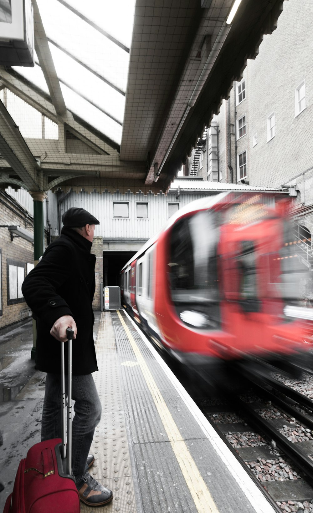 man holding luggage waiting on railway with red train passing by