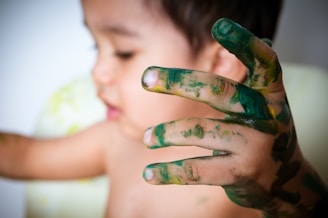 A child with their face slightly out of focus in the background, while their hand is in the foreground, covered in green, yellow, and blue paint. The child appears to be playing or experimenting with finger painting.