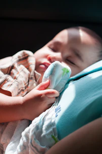 A baby is peacefully sleeping while holding a soft blanket. The soft sunlight highlights the baby’s gentle features, casting a warm glow over the scene. The baby wears a plaid shirt and rests on a smooth surface.