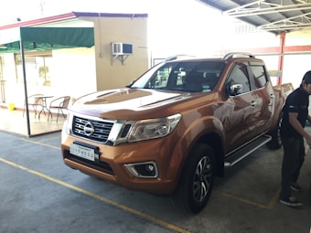 A brown pickup truck is parked in a covered area with some chairs and tables nearby. A person stands to the side of the truck, which is positioned in a parking space marked by yellow lines. There is a green awning, a building wall with an air conditioning unit, and a partial view of a garage structure.