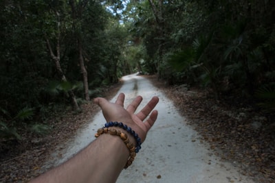 A hand wearing beaded bracelets is extended toward a narrow dirt path, lined with dense, green foliage and tall trees, creating a sense of invitation to explore or journey forward.