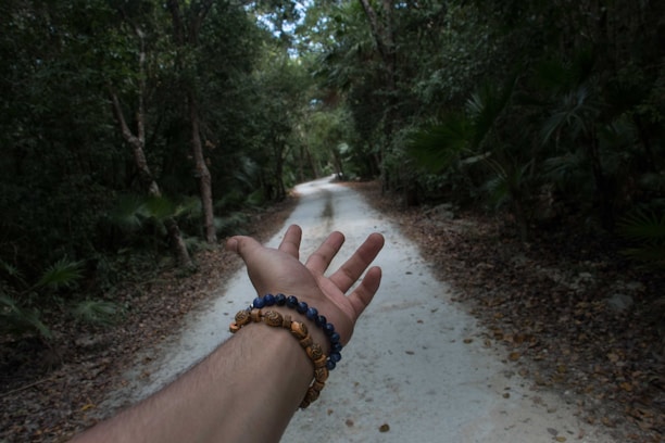 A hand wearing beaded bracelets is extended toward a narrow dirt path, lined with dense, green foliage and tall trees, creating a sense of invitation to explore or journey forward.