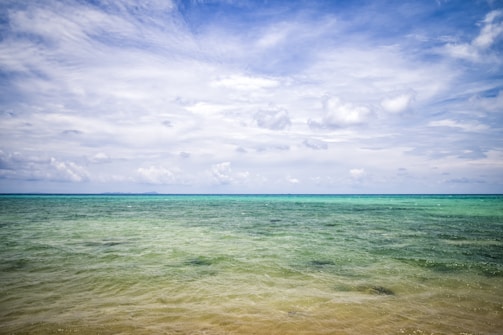 A serene seascape with a vast expanse of calm ocean waters extending towards the horizon. The sky is partly cloudy with soft, white clouds scattered across a blue sky, creating a peaceful and tranquil atmosphere.