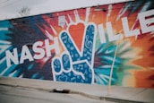 A vibrant mural on a brick wall featuring the word 'NASHVILLE' in bold, white letters. A large peace sign is incorporated into the design, filled with musical notes and swirls on a blue background. The surrounding wall is painted with dynamic, multicolored patterns, creating a lively and artistic scene.