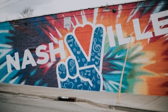 A vibrant mural on a brick wall featuring the word 'NASHVILLE' in bold, white letters. A large peace sign is incorporated into the design, filled with musical notes and swirls on a blue background. The surrounding wall is painted with dynamic, multicolored patterns, creating a lively and artistic scene.