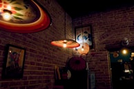 A dimly lit interior featuring exposed brick walls adorned with vintage posters and decorative lighting fixtures. The hanging lamps have colorful shades and emit a warm glow. Various decorative objects, including an umbrella and a basket lamp, add an eclectic touch to the space.